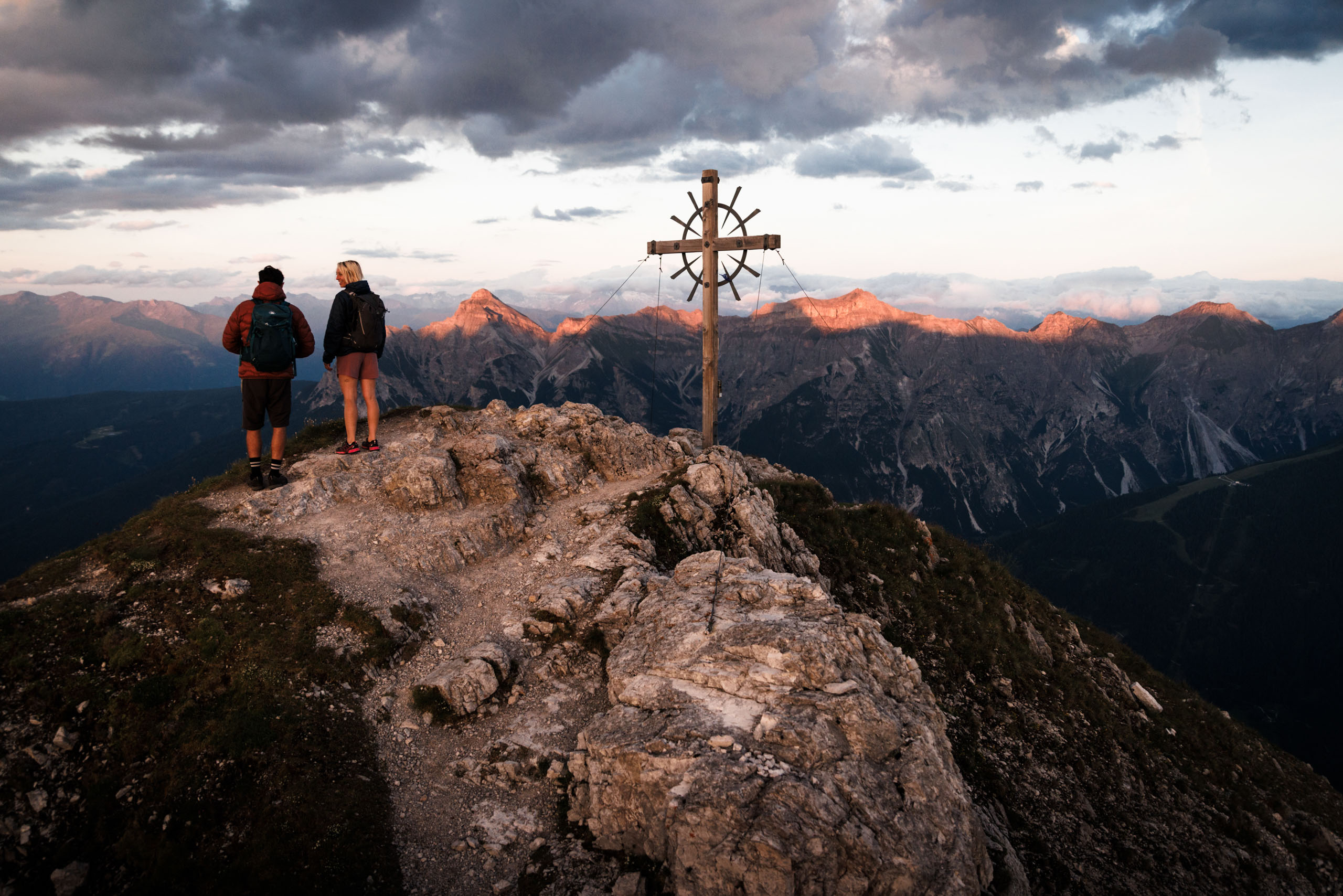 Ein Bergsteiger und eine Bergsteigerin auf dem Gipfel neben einem Gipfelkreuz bei Sonnenaufgang